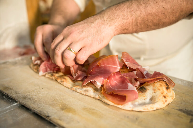mężczyzna w obrączce kładzie szynkę na cienkiej pizzy mężczyzna układa szynkę na cieście do pizzy